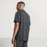 Man wearing a dark patterned short-sleeve shirt with gold stripe piping along the sleeves, viewed from the side/back.