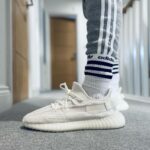 Close-up of a white athletic sneaker with striped socks and gray Adidas track pants in a hallway.