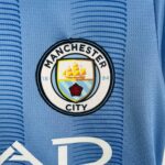 Close-up of a Manchester City crest on a blue jersey fabric, showing the shield and emblem details.