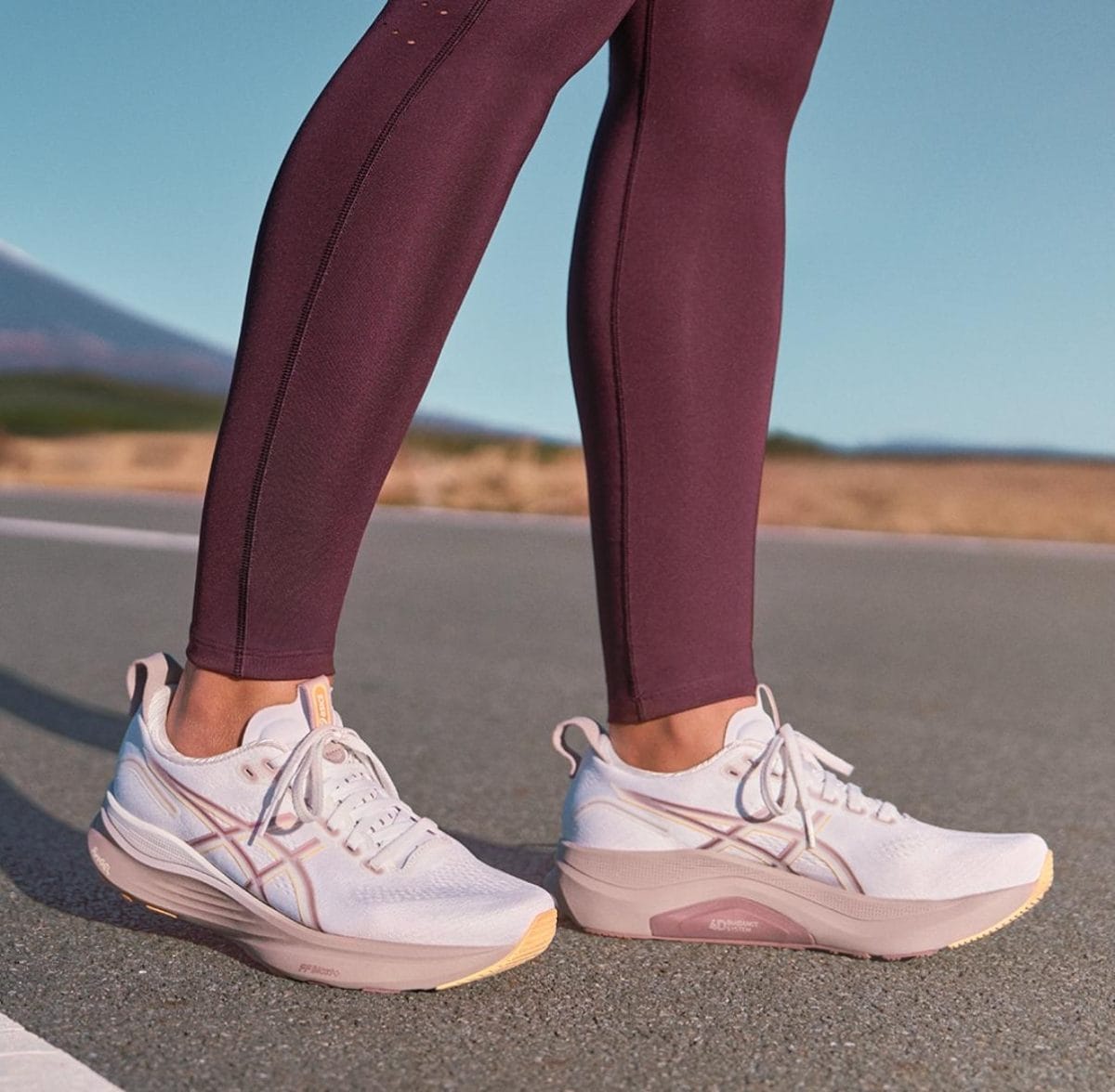 Close-up of a person wearing burgundy leggings and white pink-accent athletic shoes standing on a paved road outdoors.