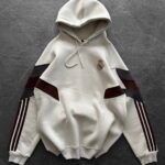 Cream hoodie with Real Madrid crest on chest and maroon-black striped sleeves laid flat on dark surface.