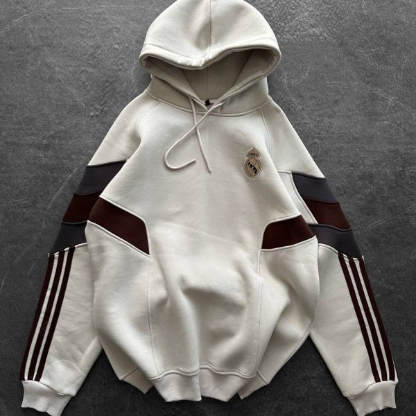 Cream hoodie with Real Madrid crest on chest and maroon-black striped sleeves laid flat on dark surface.