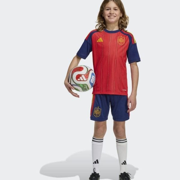 df857064-735f-4b98-ba57-bcb16b2820f2 - UAE SPORT STORE Young soccer player in a Spain kit, red shirt and blue shorts, holding a soccer ball and smiling.