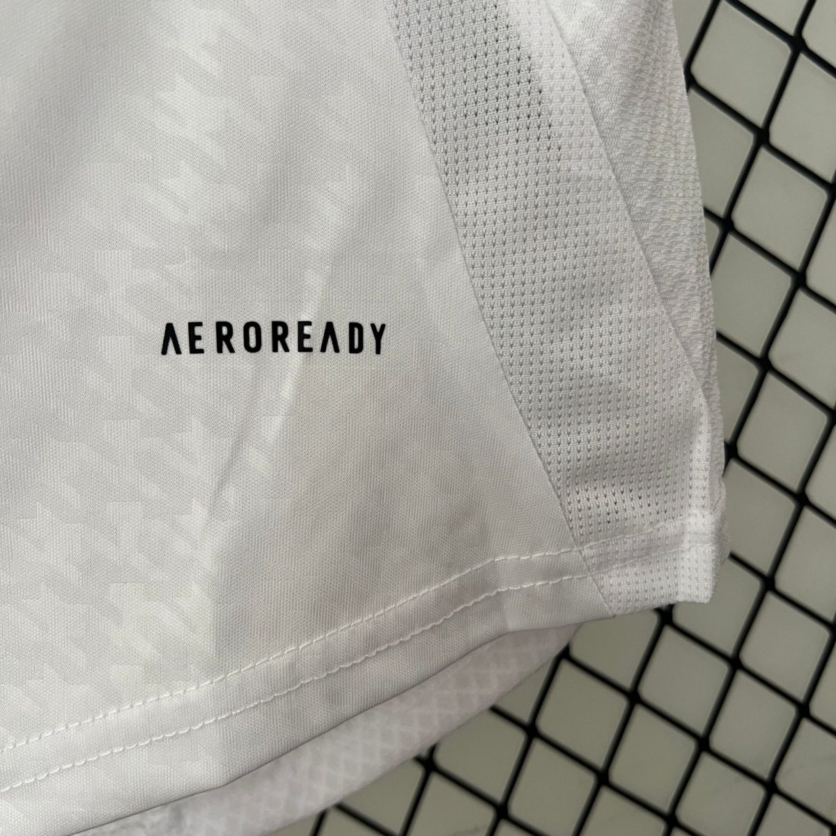 Close-up of white fabric with stitched seams and the word AEROREADY visible, set against a dark, tiled background grid.