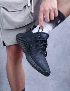 Person wearing a black athletic shoe and white sock, lifting their foot to adjust the laces against a beige short.