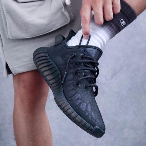 Person wearing a black athletic shoe and white sock, lifting their foot to adjust the laces against a beige short.
