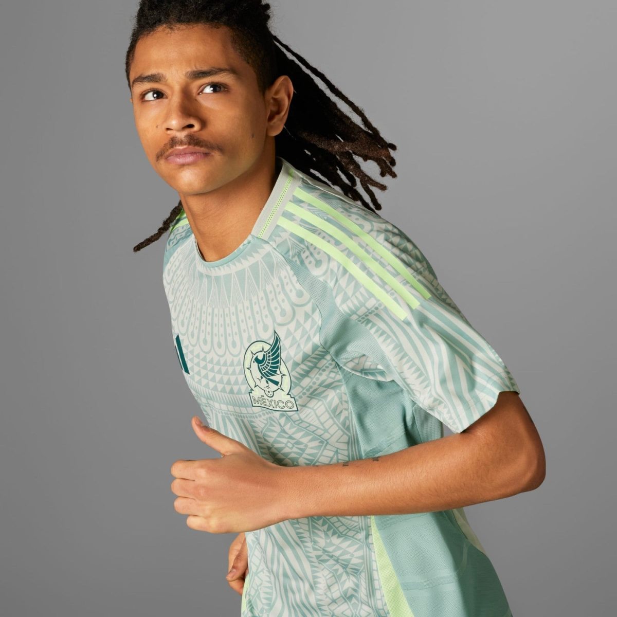 Young man with braided hair in a light green Mexico soccer jersey running or posing mid-motion against a gray backdrop, looking to the left.