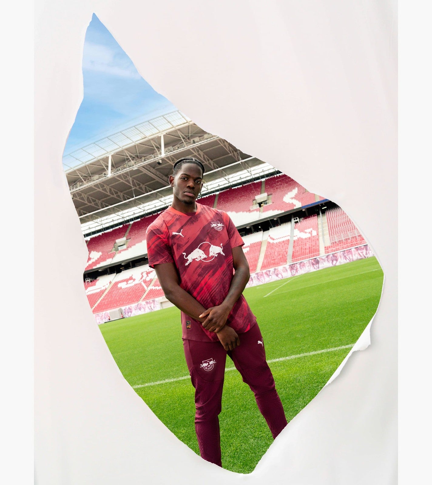 doXch3KnJVZ9ooL | UAE SPORT STORE Professional footballer in maroon kit standing on a vibrant green pitch inside a stadium, framed by a torn-paper silhouette