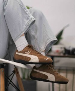 Foot wearing brown Puma sneakers with a white stripe, white socks, and light denim jeans. The person is seated with legs crossed.
