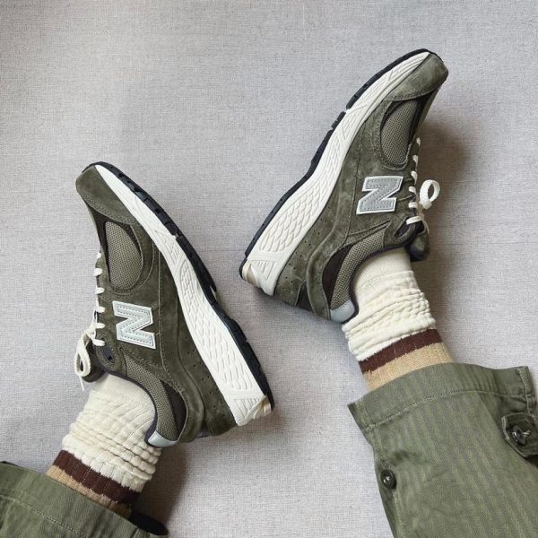 Person wearing olive-green New Balance sneakers with white soles, cream socks, and green pants standing on a light gray background.