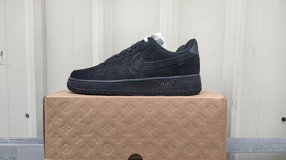 Black suede low-top sneaker with a subtle monogram pattern sits on a brown cardboard shoebox, showing the word “AIR” on the midsole edge.