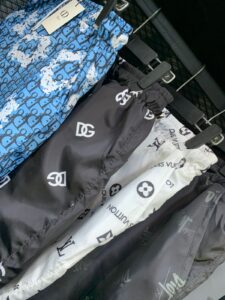 Clothing racks with designer-patterned pants, including blue patterned bottoms and black and white items, each featuring prominent luxury-brand logos (Chanel, Louis Vuitton) hanging on a metal rail.