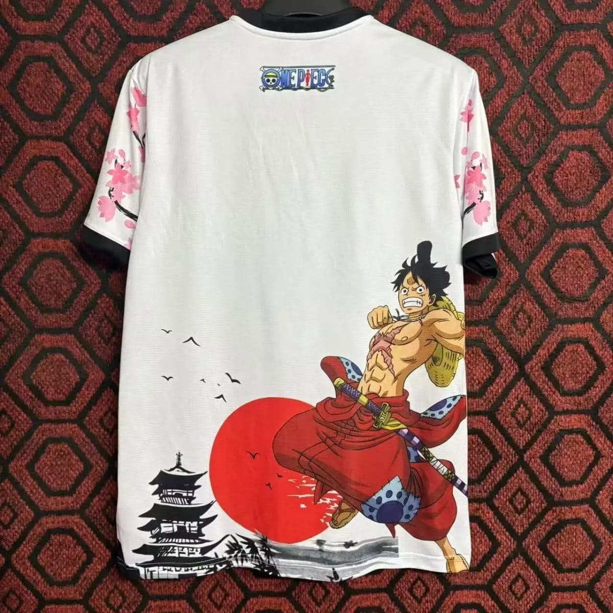 T-shirt with One Piece character Luffy battling at sunset, red sun and traditional Japanese pagoda scene on white fabric with pink blossoms on sleeves
