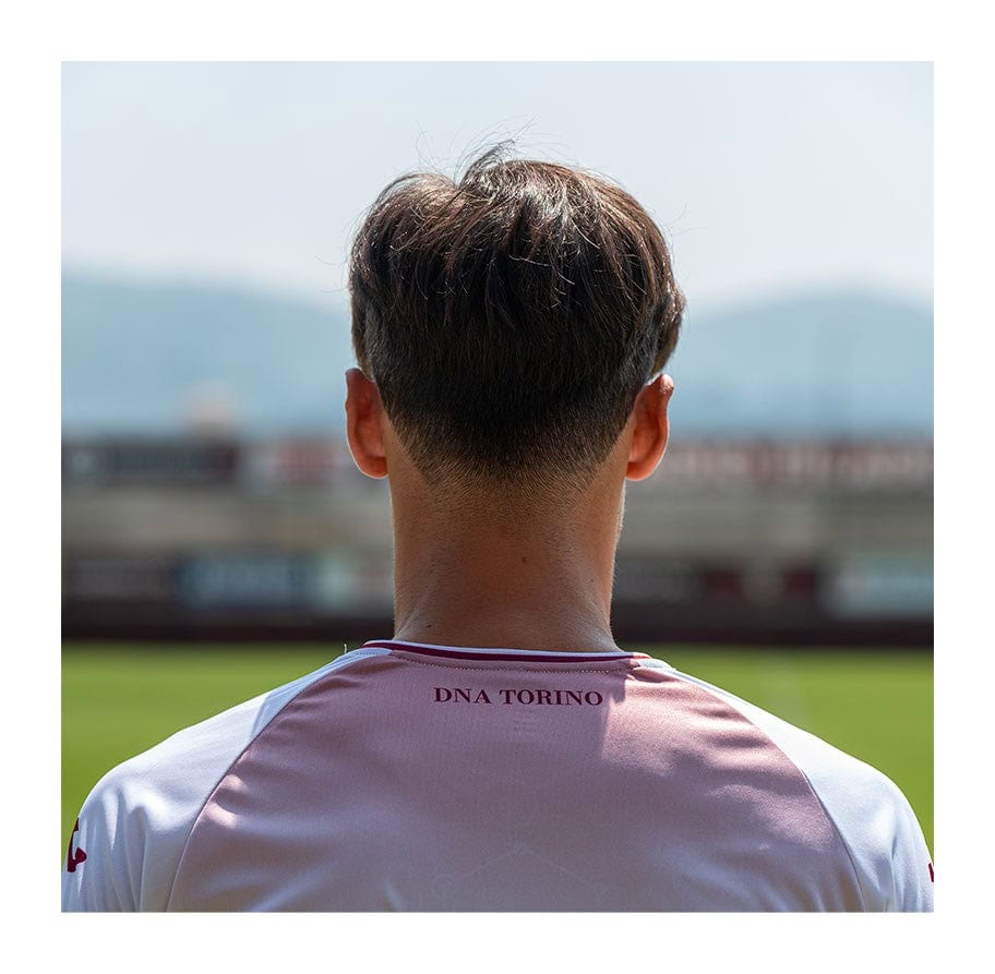 e1gZOSWsS0iPx7d | UAE SPORT STORE Back view of a person wearing a pink and white jersey with 'DNA TORINO' on the back, standing on a soccer field with a blurred stadium in the background.
