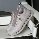 Light gray On running sneaker with a cloud-like sole resting on a white display stand in a showroom setting.