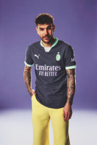 Athlete posing in a dark gray soccer jersey with mint-green trim and Emirates Fly Better sponsor, standing against a purple background with yellow pants showing tattooed arms.