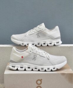 Pair of white On running shoes with chunky segmented soles displayed on a beige box against a blue-gray wall, one shoe atop the other.