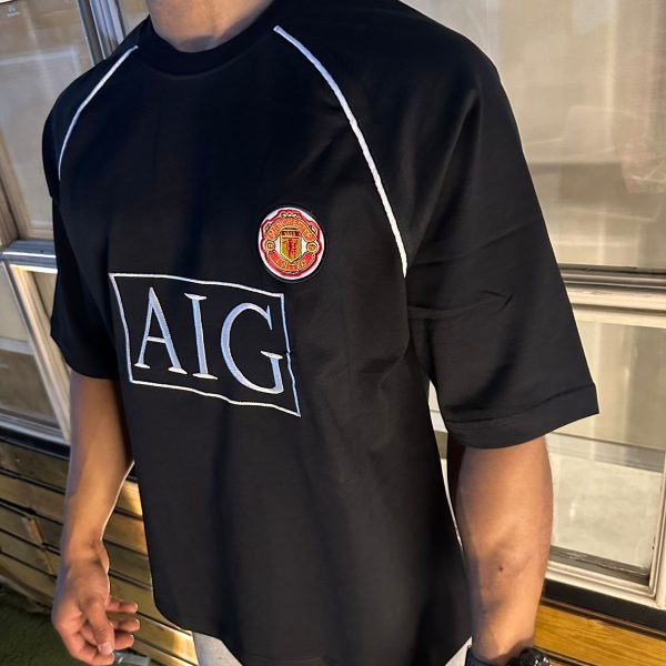 Man wearing a black Manchester United jersey with the AIG logo, standing indoors near a bench and windows.