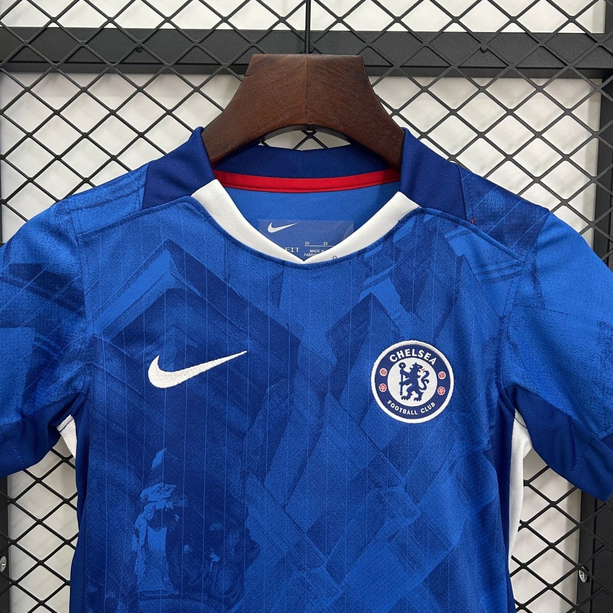 Blue Chelsea FC football jersey on a wooden hanger, crest visible on the chest against a metal grid background.