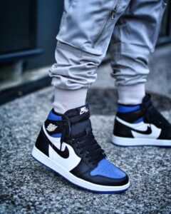 Pair of black, white, and blue Nike Air Jordan 1 sneakers worn with light gray joggers on a concrete pavement.