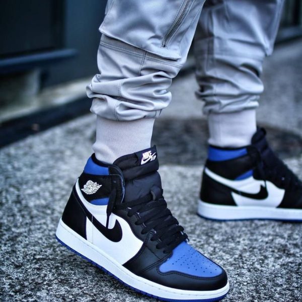 Pair of black, white, and blue Nike Air Jordan 1 sneakers worn with light gray joggers on a concrete pavement.