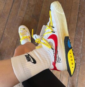 Person wearing a white sneaker with a red swoosh, bright yellow laces, and a chunky beige textured sole; white sock with a black Nike logo visible.