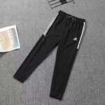 Black Adidas track pants with white side stripes and drawstring waist laid on a gray carpeted floor.