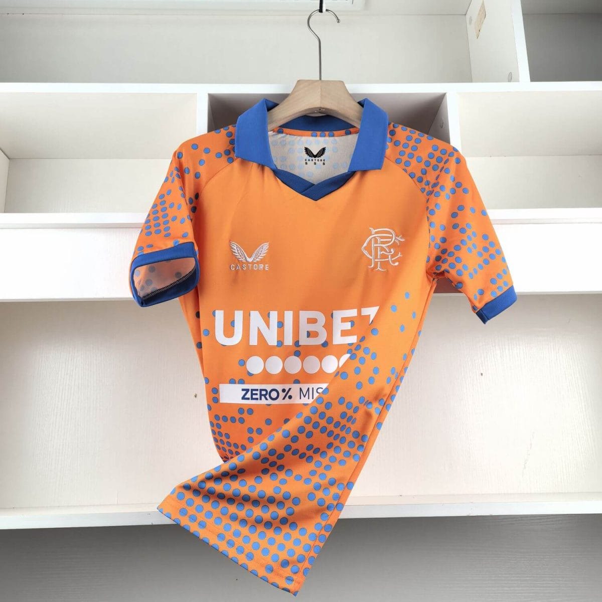 Orange sports jersey with blue polka dots hanging on a wooden hanger in a white closet; shows Castore logo and a team crest on the chest.