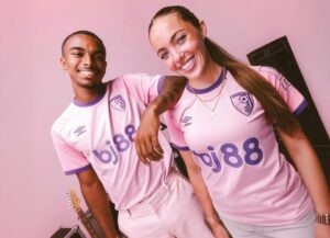 Two smiling people in pink soccer jerseys pose together in a pink room; a guitar is visible in the background.