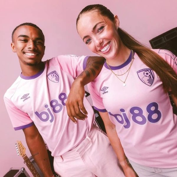 Two smiling people in pink soccer jerseys pose together in a pink room; a guitar is visible in the background.