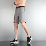 Male model wearing gray Nike athletic shorts with a black waistband and logo, paired with black-and-white sneakers in a side pose.