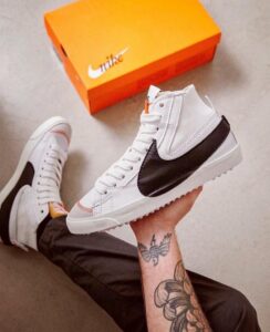 White high-top sneaker with a black Nike swoosh held over an orange shoebox on a beige carpet floor; visible tattooed forearm holding the shoe.