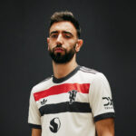 Man with short dark hair and beard in a white soccer jersey with red and black stripes, posing against a dark background.