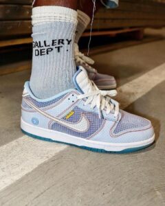 Light blue Nike sneaker with a grid pattern and white laces, worn with light blue socks reading 'GALLERY DEPT.' on a concrete floor.