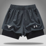 e7e8fe88 | UAE SPORT STORE Gray athletic shorts with zip pockets, layered over black compression shorts; bull head logo on left leg and Under Armour emblem on right.