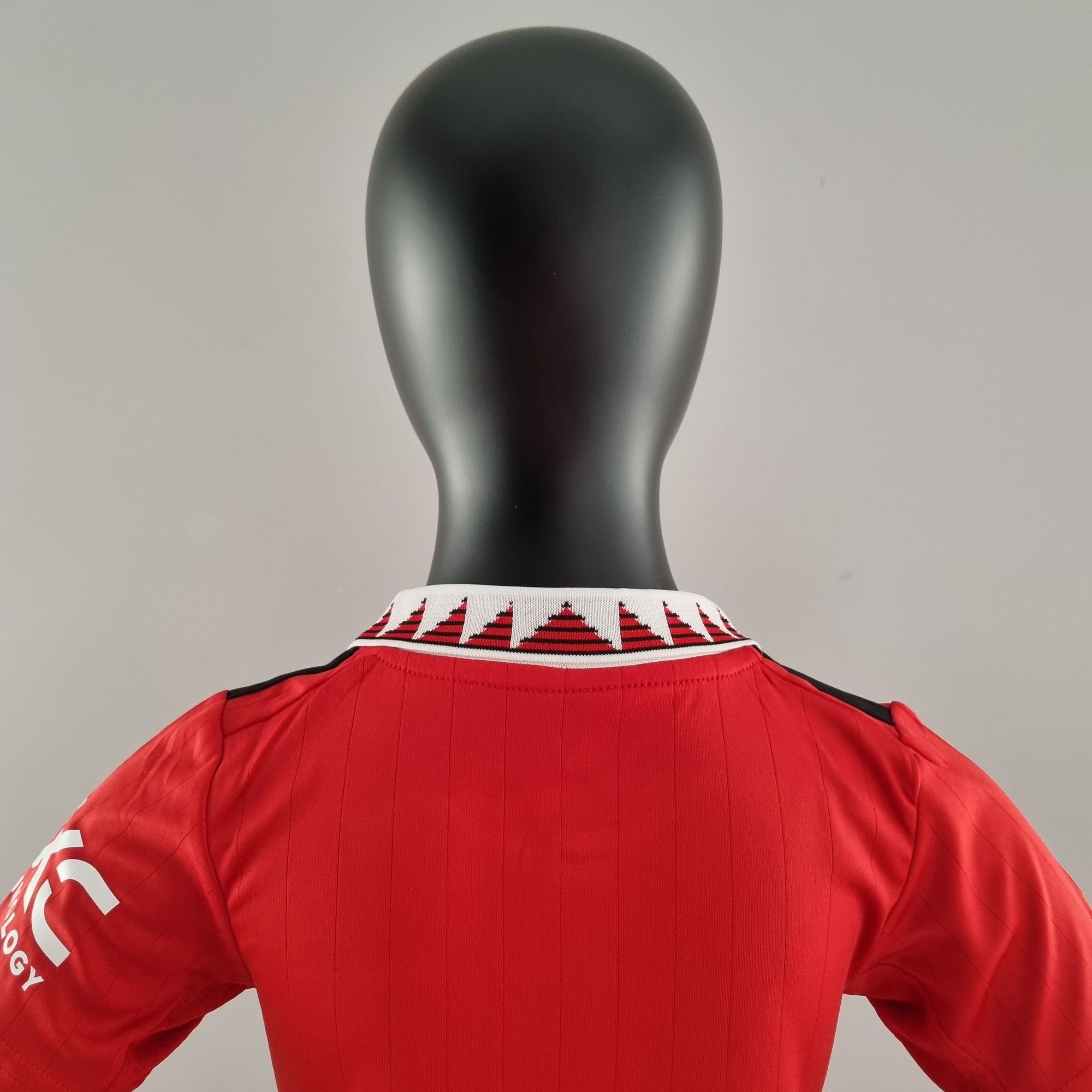 e7f632be_9303e14c-8830-4602-b5a0-5e50c16ab0ae | UAE SPORT STORE Back view of a black mannequin wearing a red athletic jersey with a white patterned collar; partial white text is visible on the sleeve (ending in 'LOGY').