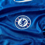 Chelsea Football Club crest on a blue jersey, circular badge with a lion holding a staff in the center, surrounded by 'Chelsea Football Club' text and small red embellishments