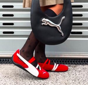 e8932bdc-2b7f-4d24-9052-c5773f3b8efc | UAE SPORT STORE Person wearing red and white Puma sneakers, black patterned tights, and carrying a black handbag with a silver Puma logo against a metal wall backdrop.