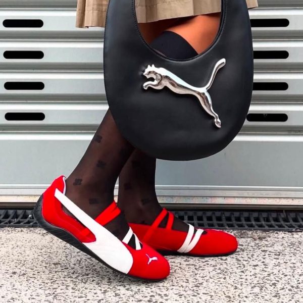 Person wearing red and white Puma sneakers, black patterned tights, and carrying a black handbag with a silver Puma logo against a metal wall backdrop.