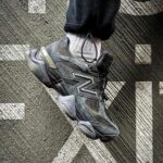 Close-up of a dark gray New Balance-style sneaker with a bold side logo, laces tied, worn with a light gray sock, resting on a textured, patterned carpet.