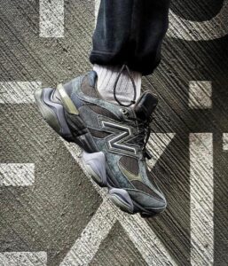 e8b95b1a-ab1e-4c5c-9fda-93eb9564c313 | UAE SPORT STORE Close-up of a dark gray New Balance-style sneaker with a bold side logo, laces tied, worn with a light gray sock, resting on a textured, patterned carpet.