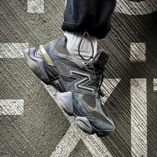 Close-up of a dark gray New Balance-style sneaker with a bold side logo, laces tied, worn with a light gray sock, resting on a textured, patterned carpet.