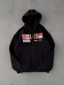 Black zip-up hoodie with racing sponsor patches on the chest, hood up, set against a gray concrete wall.