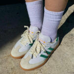 Person wearing light lavender socks and white sneakers with green stripes, standing on a concrete surface with tan soles.