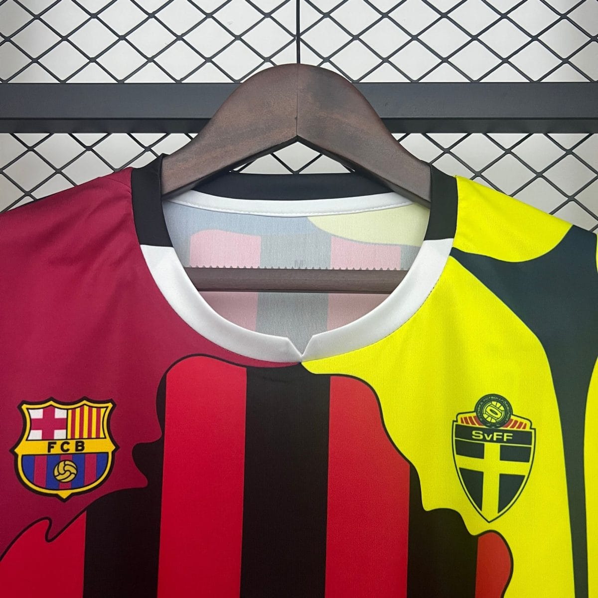 Close-up of two football jerseys on a wooden hanger: Barcelona crest on the left and a second crest on the right, against a chain-link fence backdrop.