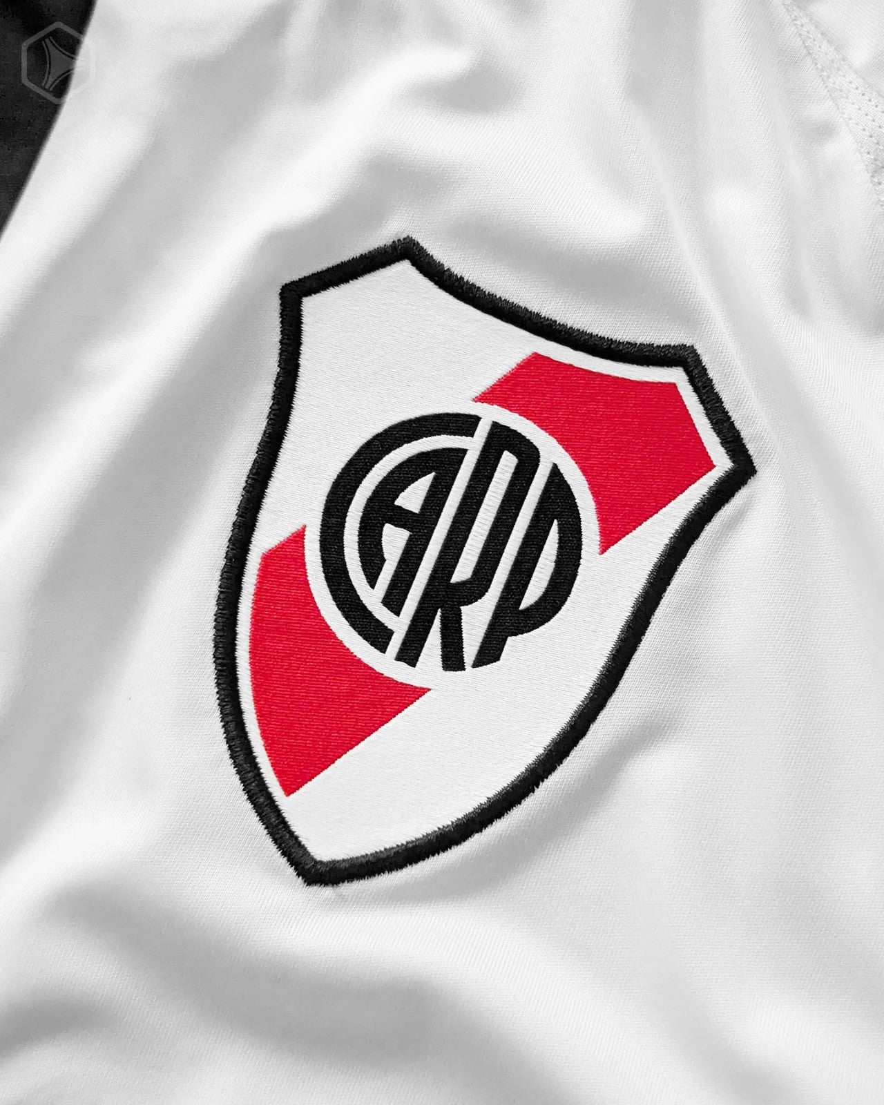 eBRsnHQZYcHtdnD | UAE SPORT STORE White fabric featuring an embroidered shield crest with a red diagonal stripe and a black border surrounding a circular monogram.
