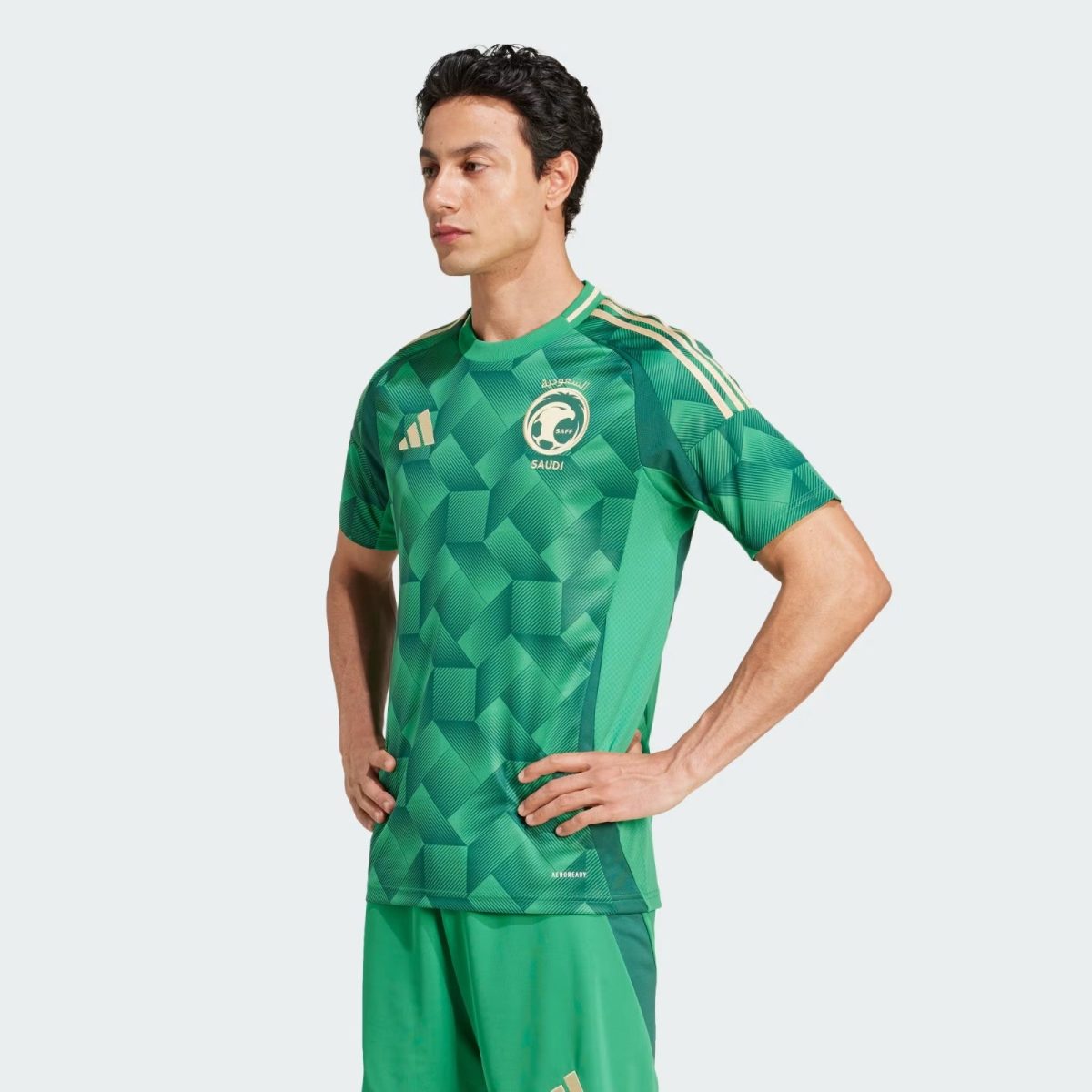 Male model wearing a green Saudi national football jersey with a geometric pattern and Adidas stripes across the shoulders, standing with hands on hips.