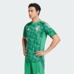 Male model wearing a green Saudi national football jersey with a geometric pattern and Adidas stripes across the shoulders, standing with hands on hips.
