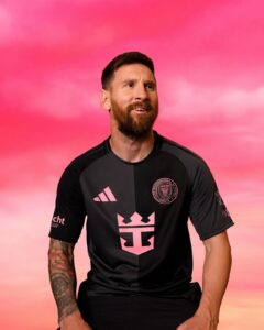 Man wearing a black athletic jersey with pink logos and a crest stands against a pink sunset sky, looking to the side.
