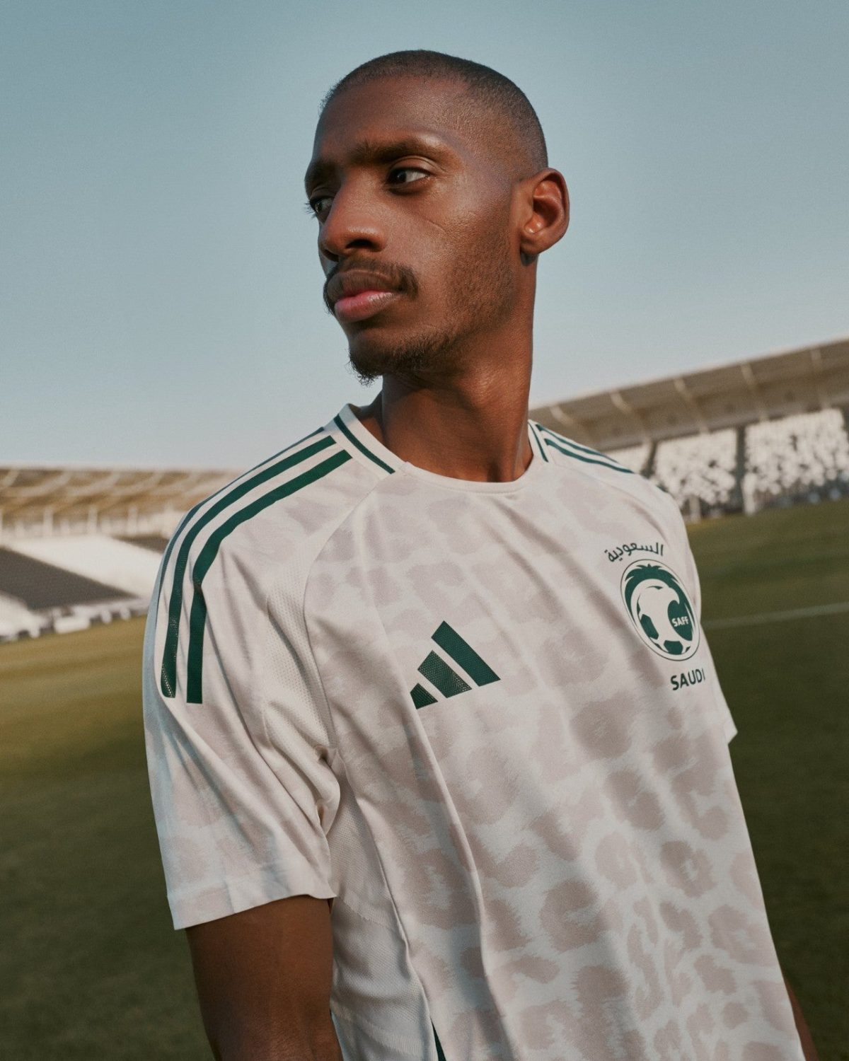 Portrait of a man in a white Saudi football jersey standing on a grassy field with a stadium in the background, looking off to the side.